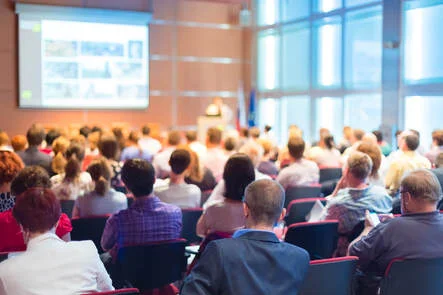 A imagem é uma foto de uma palestra, com uma apresentação e uma plateia com bastante gente. Trata-se de uma atividade comumente observada em eventos profissionais