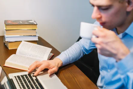 A imagem representa o fenômeno publish or perish (publique ou padeça), contendo a imagem de um pesquisador com semblante tenso sentado em uma mesa de trabalho, digitando em um laptop com uma mão e na outra uma xícara de café. No cenário há um livro aberto e outra pilha de livros, conferindo um ar de grande pressão e cobrança, inerentes ao publish or perish a que os pesquisadores são submetidos.