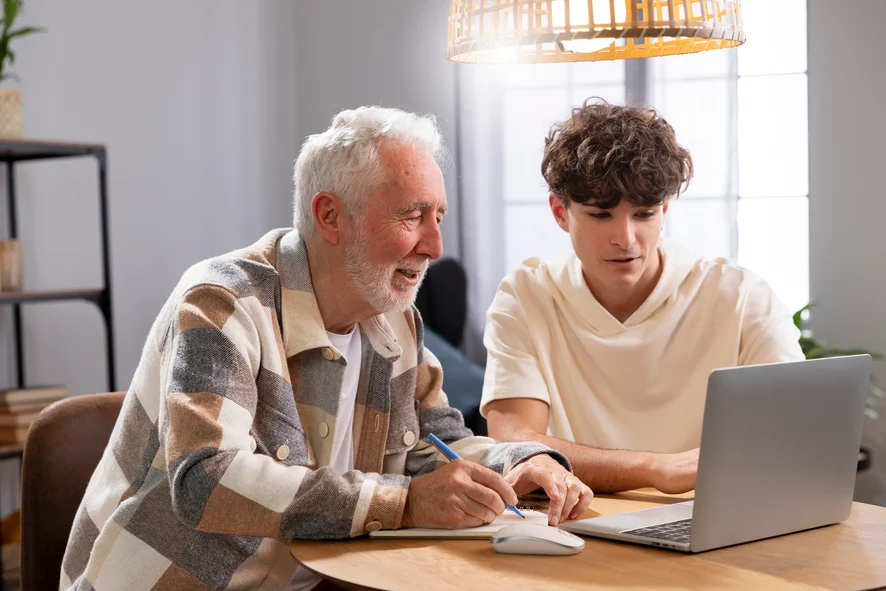 Trata-se de uma foto de um senhor com um jovem, sentados em uma mesa, na frente de um computador, estudando juntos. A aprendizagem é ao longo da vida.