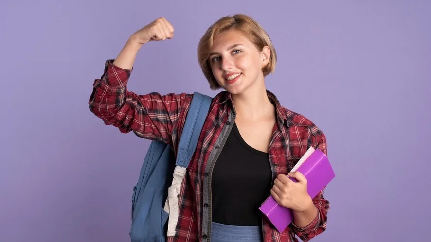 Mulher jovem segurando livros em uma mão e, no outro braço, uma alça da mochila pendurada e seu punho cerrado. Isonomia de gênero é necessário no em todos os espaços, especialmente no ambiente universitário