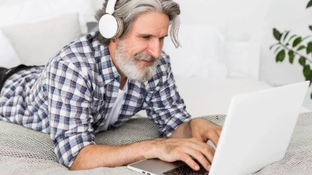 homem grisalho deitado na cama com um laptop aberto e fone no ouvido, estudando e apredendo com autonomia baseado na educação híbrida 4.0