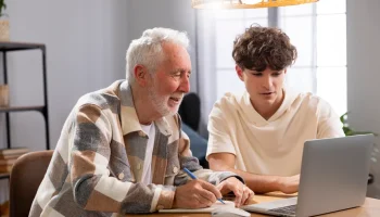 Trata-se de uma foto de um senhor com um jovem, sentados em uma mesa, na frente de um computador, estudando juntos. A aprendizagem é ao longo da vida.