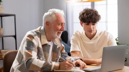 Trata-se de uma foto de um senhor com um jovem, sentados em uma mesa, na frente de um computador, estudando juntos. A aprendizagem é ao longo da vida.