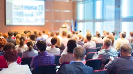 A imagem é uma foto de uma palestra, com uma apresentação e uma plateia com bastante gente. Trata-se de uma atividade comumente observada em eventos profissionais