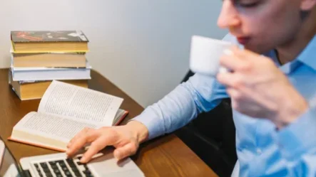 A imagem representa o fenômeno publish or perish (publique ou padeça), contendo a imagem de um pesquisador com semblante tenso sentado em uma mesa de trabalho, digitando em um laptop com uma mão e na outra uma xícara de café. No cenário há um livro aberto e outra pilha de livros, conferindo um ar de grande pressão e cobrança, inerentes ao publish or perish a que os pesquisadores são submetidos.